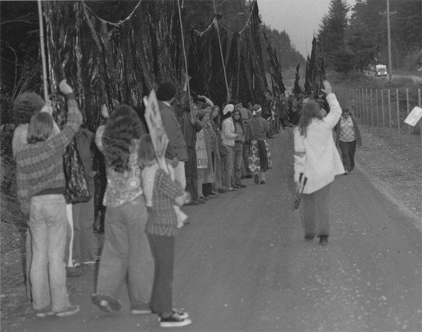 After cutting through the fence at the Trident naval base, peace activists take the “Trident Monster” inside the base.  Shelley Douglass is on the right, walking away from the camera, holding the bolt cutters.  Several children helped carry the monster over the fence.