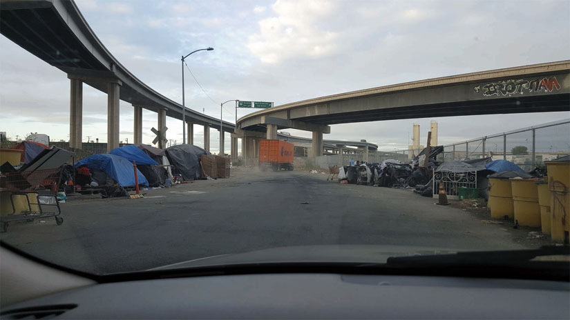 A homeless encampment under Oakland freeways, viewed through a car’s windshield. Carts, tents and belongings are clustered on the left and right. Kwalin Kimaathi photo