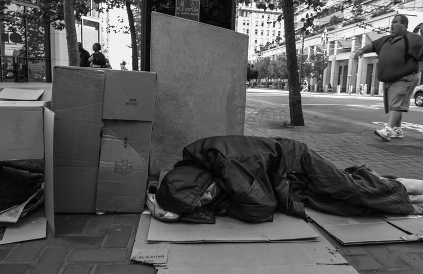 A homeless man sleeps on a San Francisco sidewalk as passers-by ignore him and pretend he’s not even there. David Bacon photo