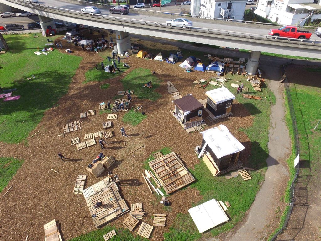 An arial view of the encampment being built. Three tiny homes and several tents stand together on a plot of land. Around them, wood pallets sit where new tiny homes will be built.