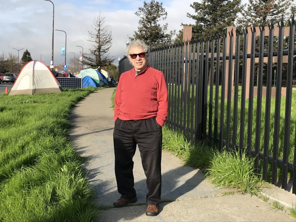 Steve Gillman poses for a photo with the Here/There encampment, and part of the "Here" sculpture, behind him.