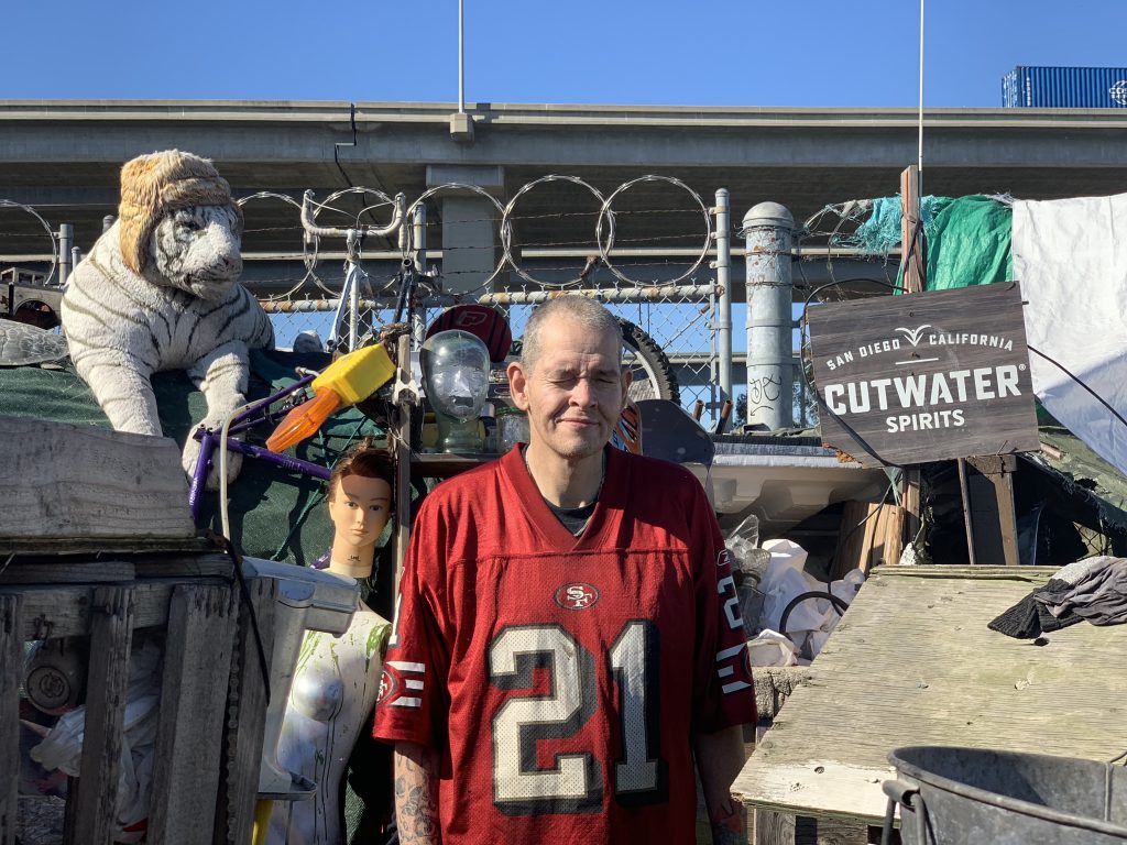 Tim Nishibori standing outside of the home that he built for himself. Surrounding him are wooden structures, a chain link fence, a mannequin doll, and a stuffed tiger.