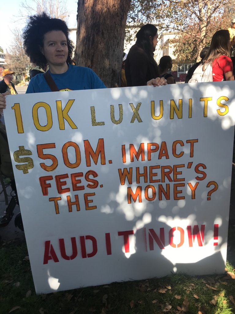 Gloria Bruce holds sign that reads, "10K Lux unites. $50 m impact fees. Where's the money? Audit now!"