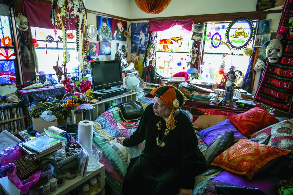 Julia Vinograd sits on her bed in a very colorful and cluttered bedroom. Her bedroom looks like a rainbow: decorative pillows and a colorful bedspread adorn her bed. Mobiles and masks hang from the ceiling and windows.  