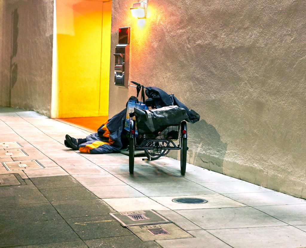 A homeless person sits on an empty Berkeley sidewalk. Only their feet are visible behind their cart.
