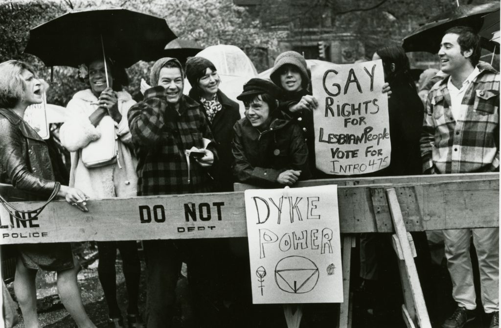 A black and white photo of a group of people at a demonstration at City Hall, New York, 1973. They are holding umbrellas. A few people hold signs that read "Dyke Power" and "Gay rights for lesbian people vote for intro 475."