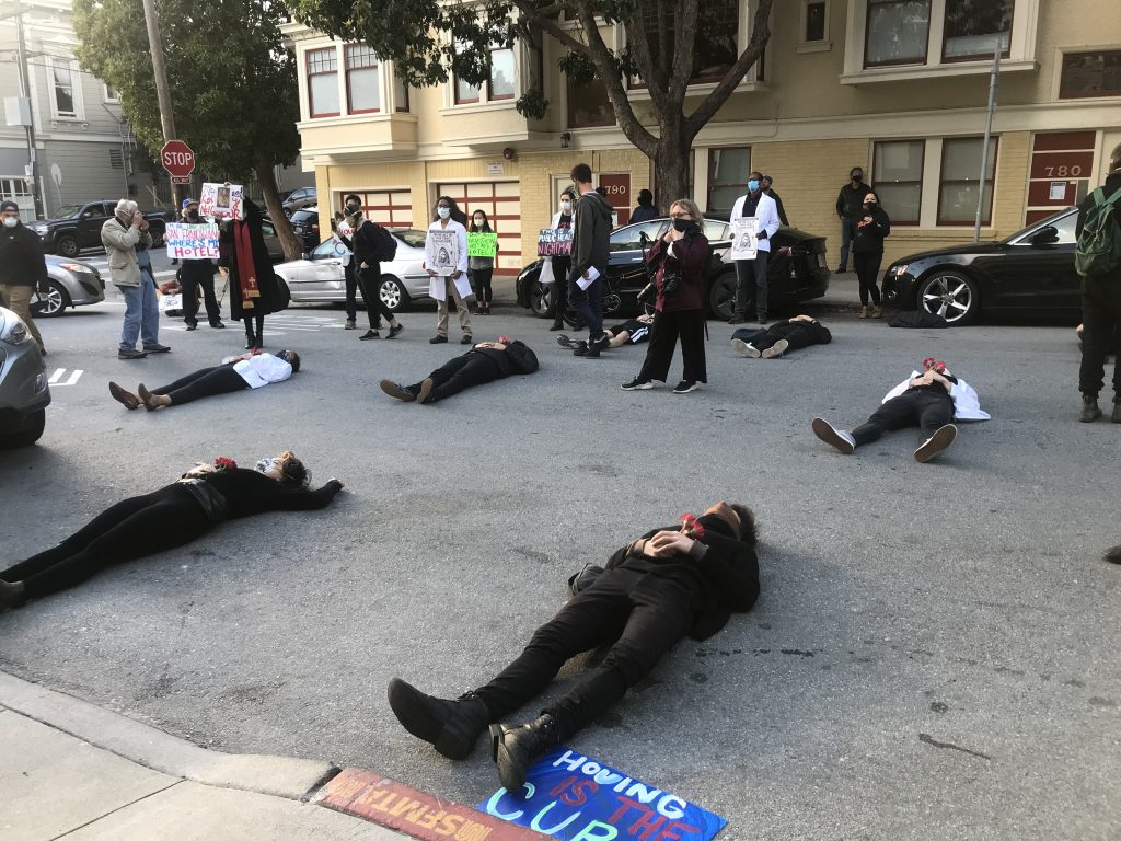 Protestors lie on the street in front of the mayor's house in San Francisco. They are wearing all black and holding red flowers.