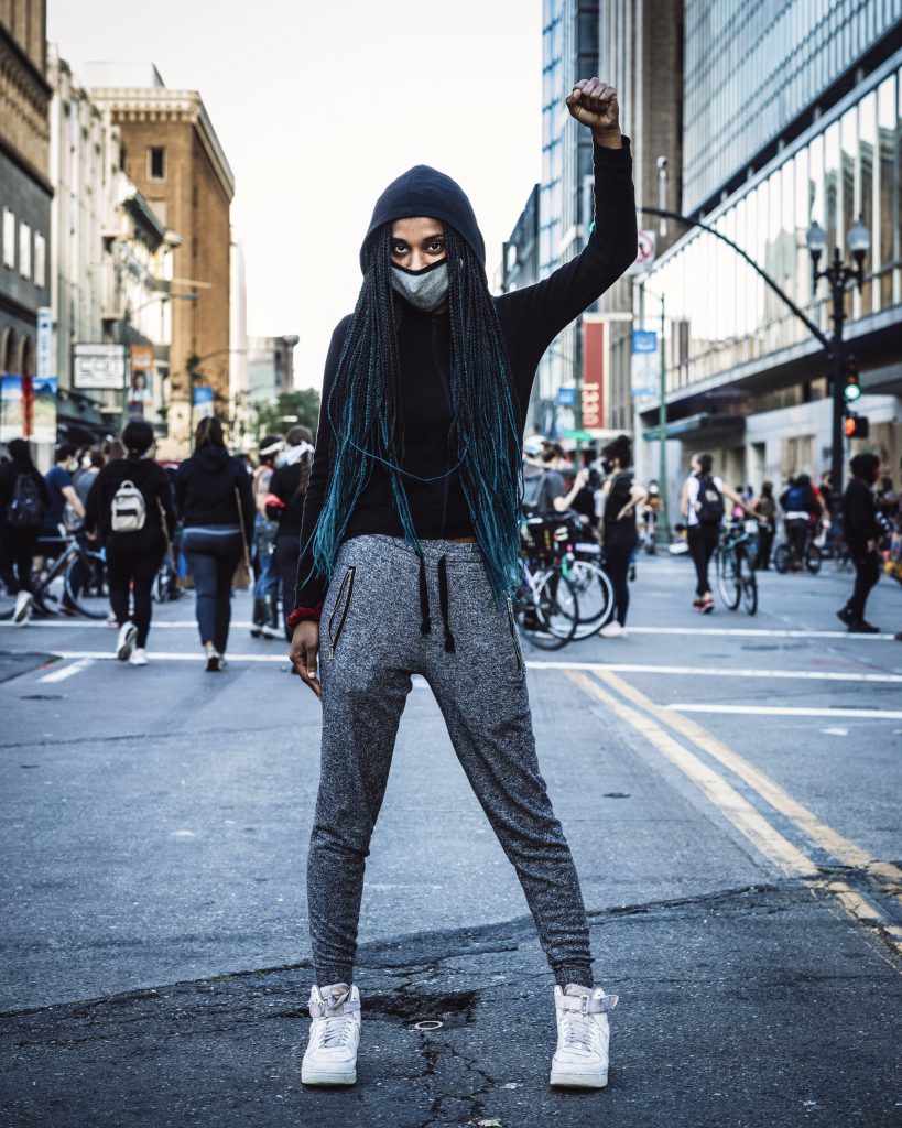 A Black woman—perhaps in her mid-20s—stands in the middle of the street in Downtown Oakland. She is looking at the photographer and holding up the fist of Black power. She has long braids which are blue on the ends.