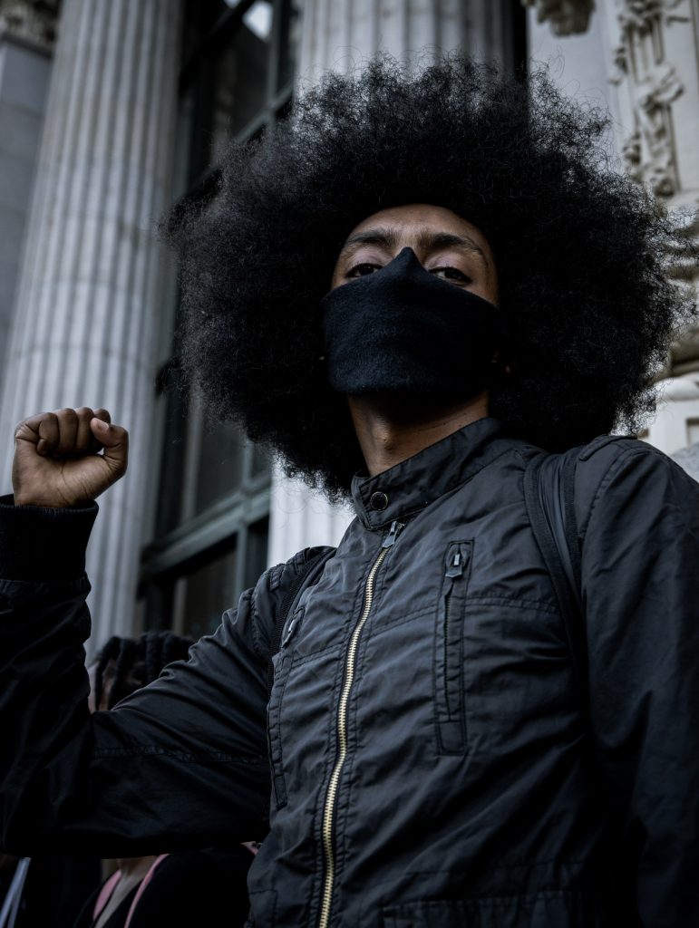 A Black man wearing a large afro and a black face mask holds up the fist of black power in front of Oakland City Hall. 