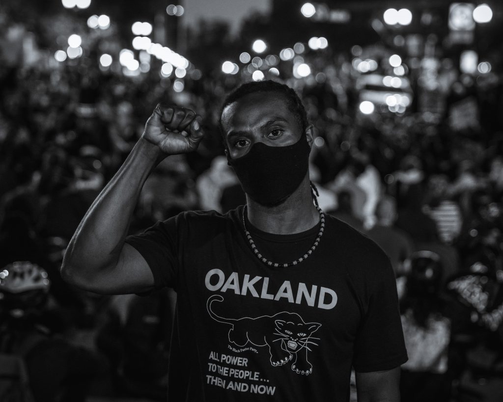 A Black man holds up the fist of black power in front of a blur of protestors and light behind him. The image is in black and white. He is wearing a shirt that says "Oakland" on it, and has a photo of a black panther. Underneath, it says "all power to the people...then and now"