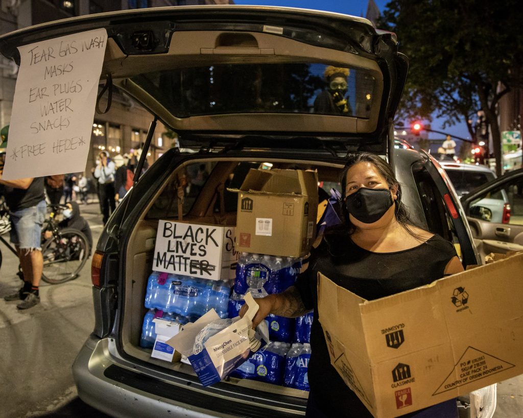 A woman wearing a face mask stands by the trunk of her car, which is open. Her trunk is full of water bottles. A sign on her open trunk reads "tear gash wash, masks, ear plugs, water, snacks, free here." A sign in her trunk reads "Black lives matter."