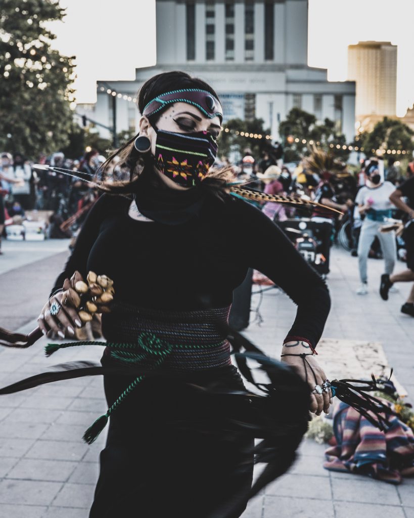 Action shot of a woman in traditional Aztec clothing dancing in front of Oakland City Hall. She is wearing all black, and there are a few bright pops of color in her outfit. She is holding a shaker in her hand and looking downward. 