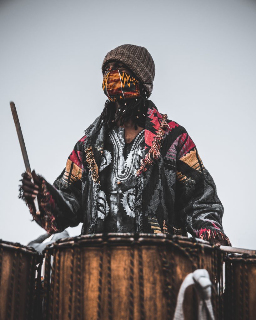 A Black drummer in wearing colorful tribal patterns and a colorful tribal face mask holds up one drum stick, as if about to hit his drum. He looks off into the distance. 