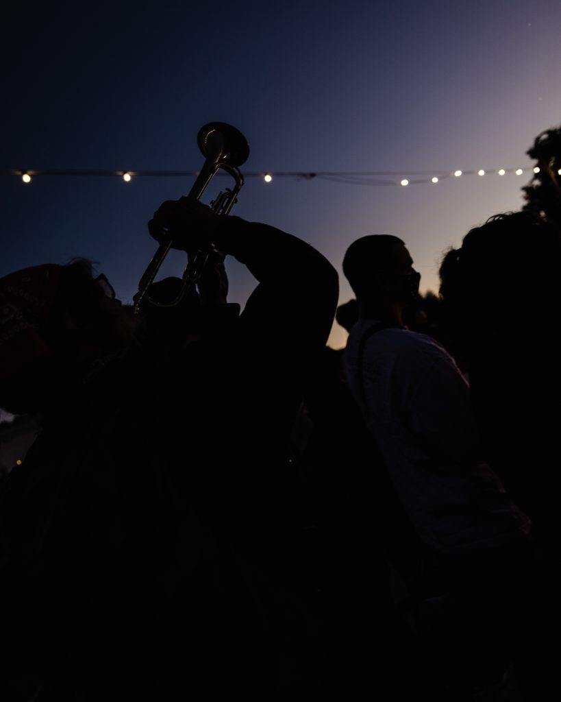 A night time image showcasing the silhouette of a trumpet player in the foreground, playing his trumpet up into the air. Behind him you can see the string lights of Lake Merritt, and the last rays of the sunset.