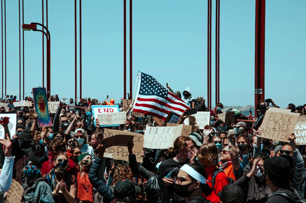 Protesters raise the American flag on the Golden Gate Bridge. Tons of people fill the frame, holding signs and standing on the roadway.