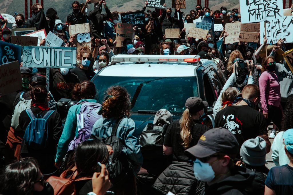 A dense throng of protestors encircle a police car on the Golden Gate Bridge. In the background, the bridge's red cables can be seen.