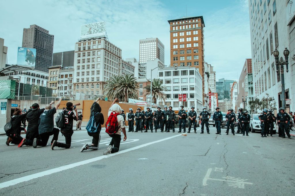 In the background, police stand in a line, blocking off Geary Street. In the foreground, several protestors make a line facing them, kneeling down with their fists in the air.