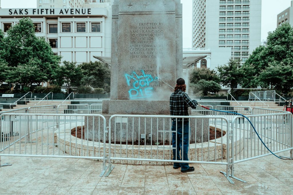 A man with a power washer removes anti-police graffiti from Union Square’s Dewey Monument. The graffiti says "all cops die" in blue spray paint.