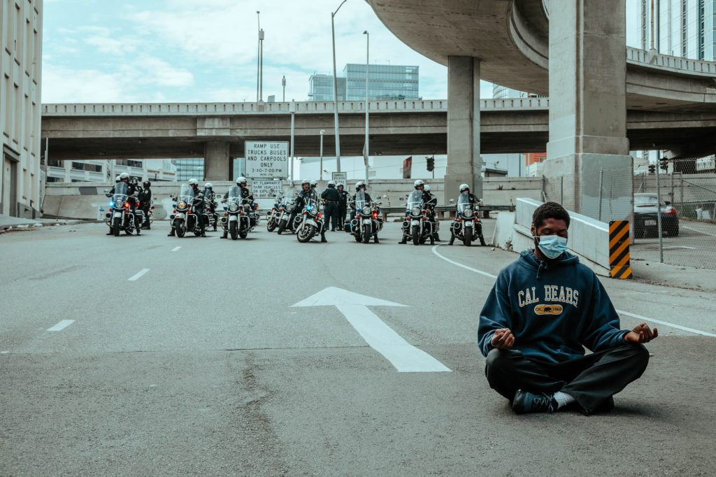 In the foreground, a protester sits on the onramp to the Bay Bridge. He is cross legged in with his hands on his knees and his eyes closed. Behind him, a line of police of motorcycles keep protesters from marching onto the Bay Bridge.
