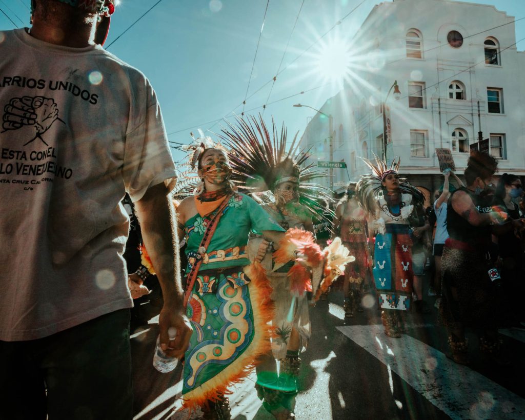 Protestors and Aztec dancers in colorful apparel and feather head dresses march toward the Mission police station.