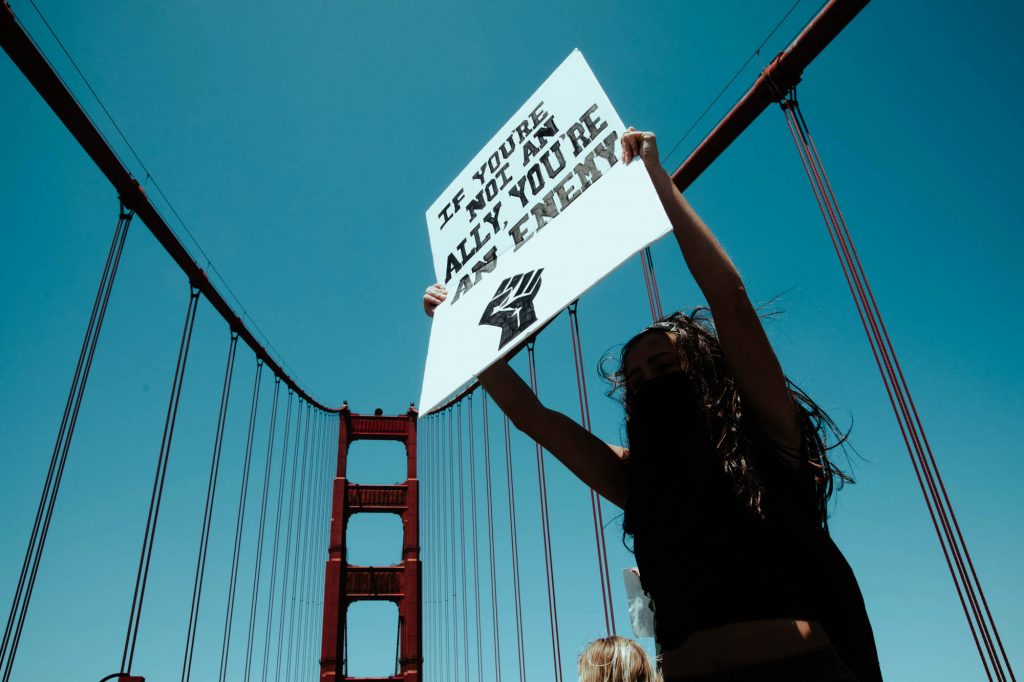 A protester holds a sign that says "if you are not an ally you're an enemy" with the black power fist below it. Behind her, the blue sky and a one of the classic bridge towers can be seen.
