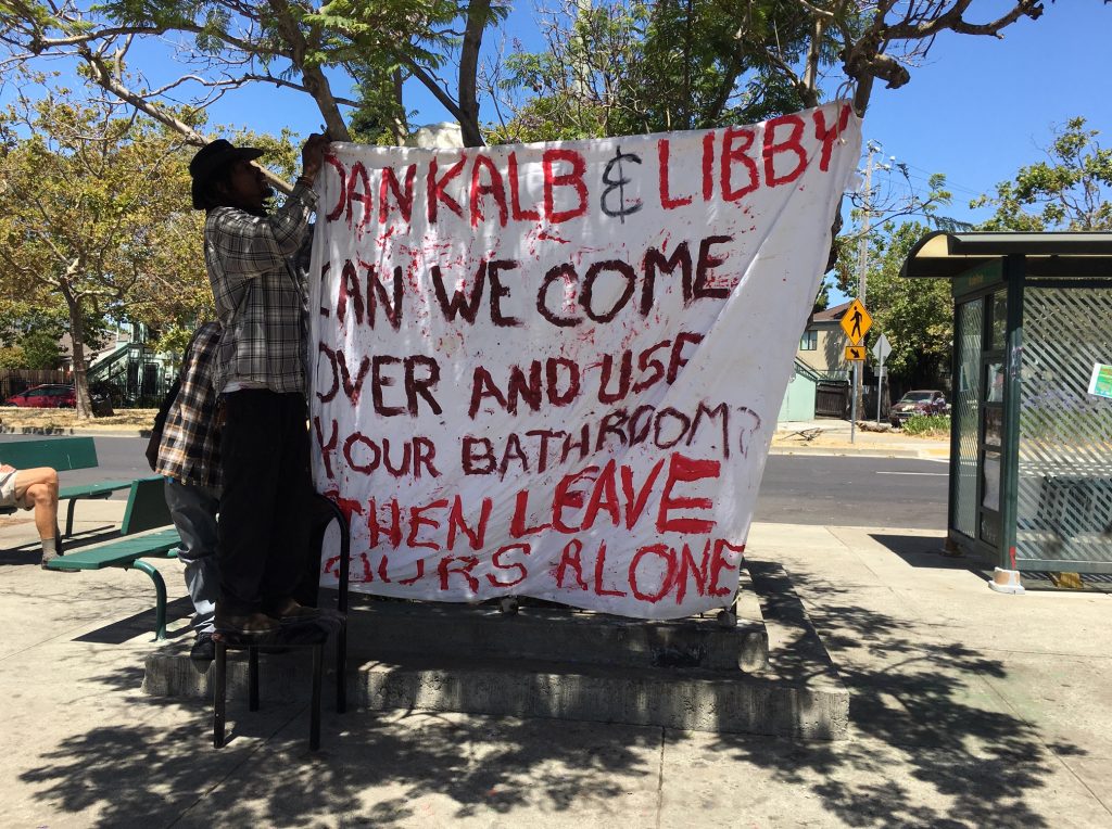 Protestors hang a banner that says "Dan Kalb & Libby, can we come over and use your bathroom? Then leave ours alone."