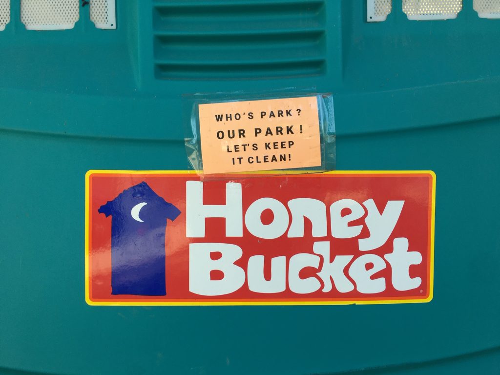 A closeup of a "honey bucket" porta potty with a sticker on it that says "who's park? Our park! lets keep it clean!"