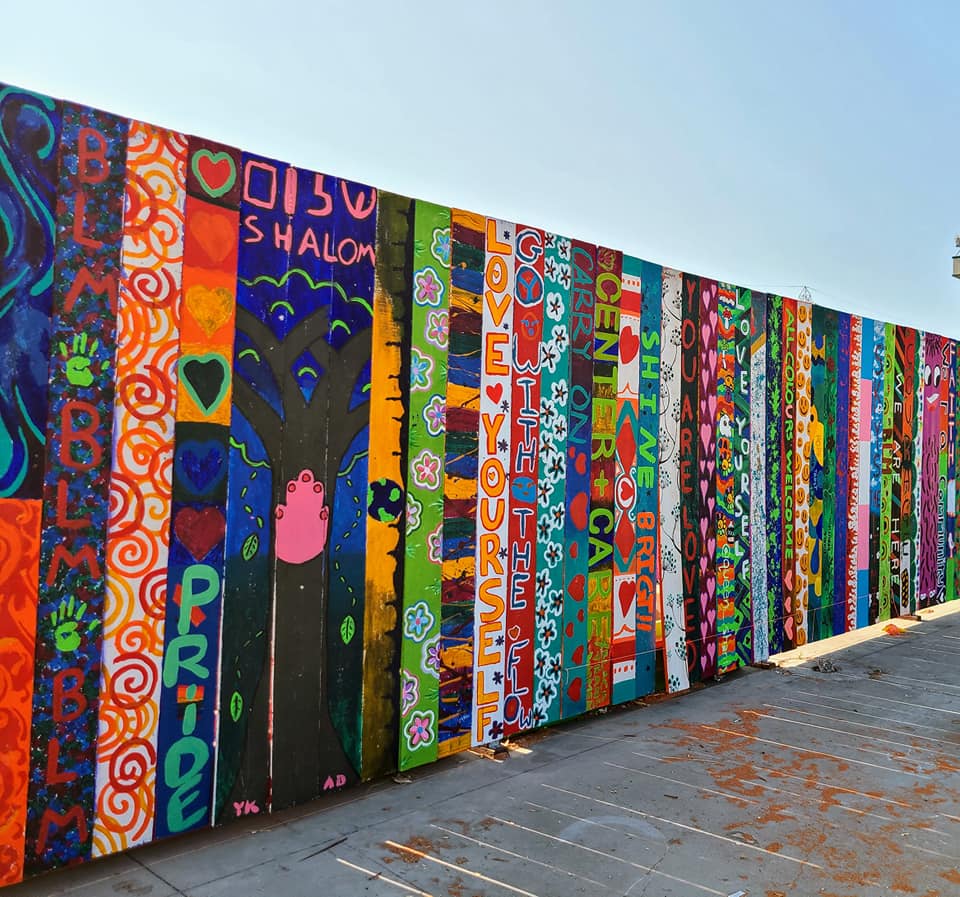 A photo of a wooden fence, Each plank of the fence has been painted by a different artist. They are colorful and contain images and writing.