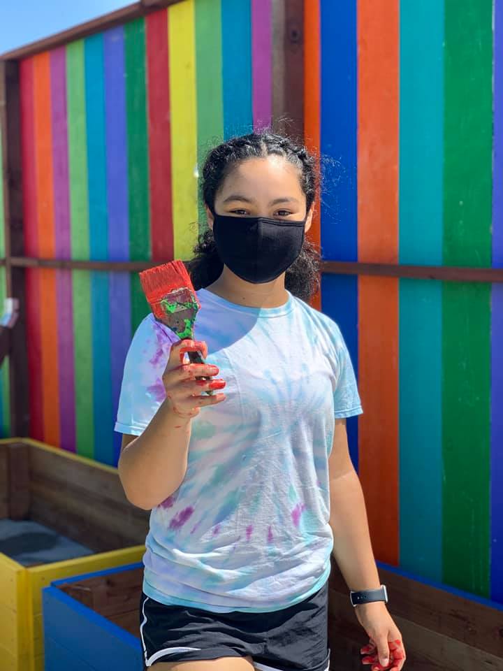A young painter stands in front of a rainbow fence with a paintbrush in her hand.