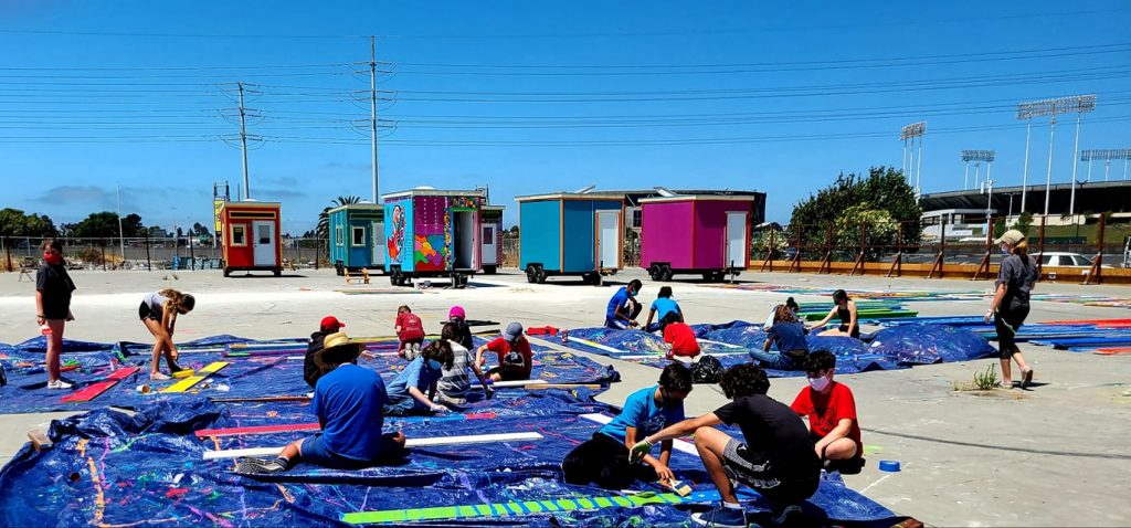 In the foreground, young people painting fence planks. In the background, six colorful tiny homes.