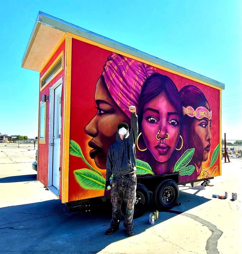 An artist stands in front of a beautiful mural on a tiny home with his fist up in the air in celebration. The mural is pink and red, of three women.