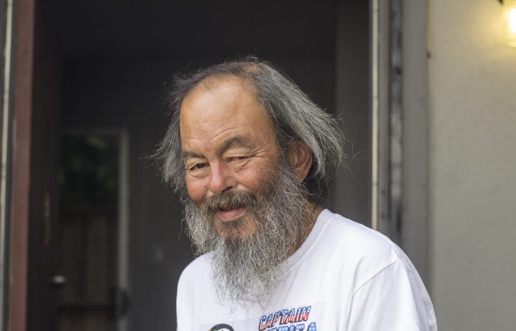 Mike Lee smiles for a photo after obtaining housing in 2017. He has a long grey beard and is wearing a white shirt.