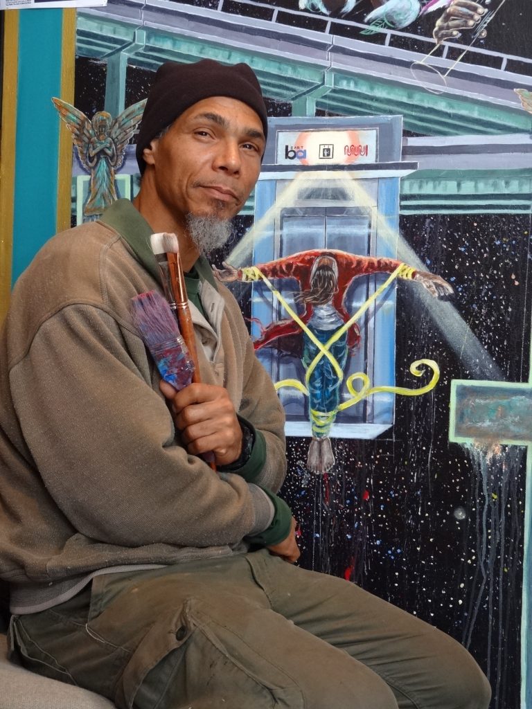 Ronnie Goodman sits in front of a painting he is working on with a paint brush in his hand.