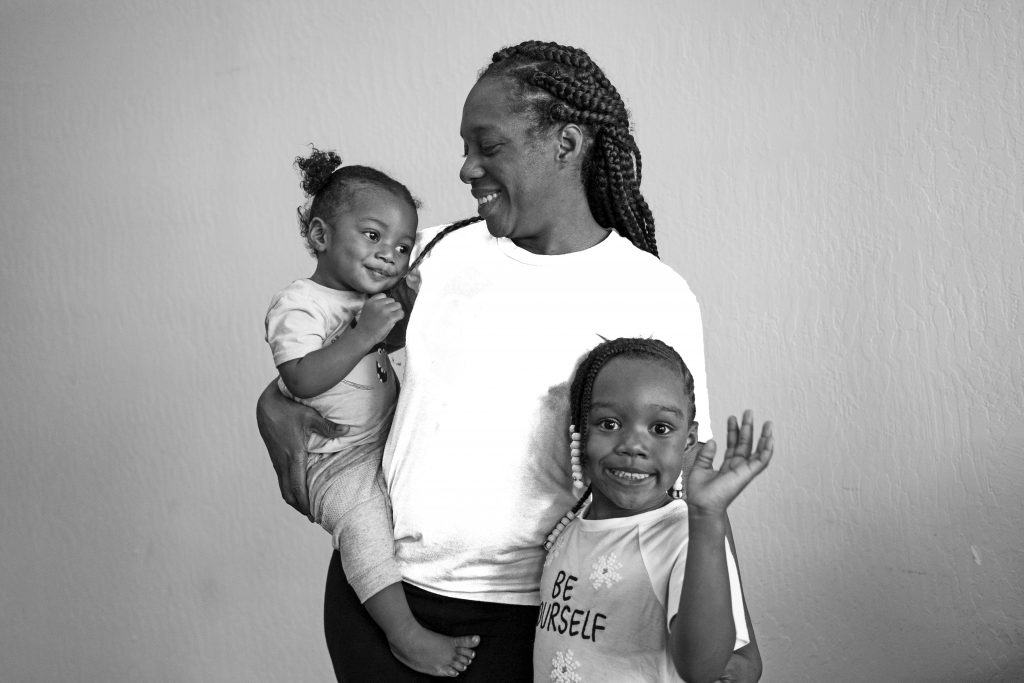 Black and white photo of Dominique and her two young children. She holds one of them in her arms as the other one stands on the group and waves at the camera. 