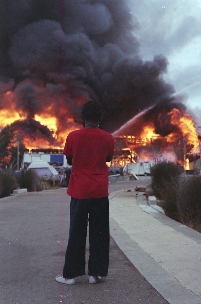 A woman with her back facing the camera watches a building engulfed in flame burn before her. She is wearing a red t-shirt, black pants, and socks. Her arms are crossed as she observes the building go up in dramatic red and orange flames.