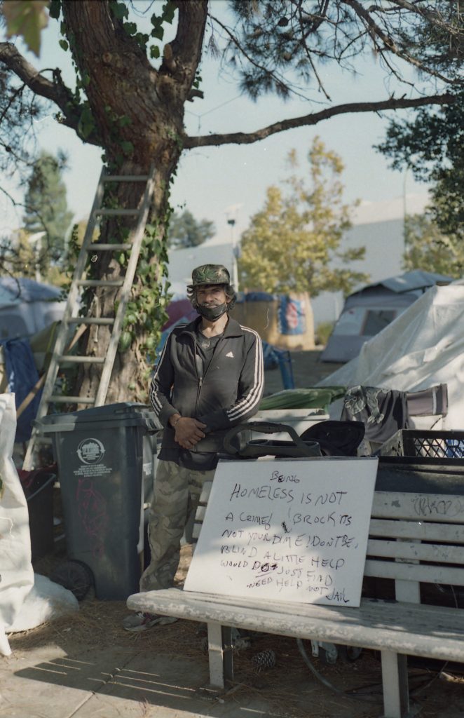 An encampment residents stands next to a tree. In front of him, a sign he made reads, "Being homeless is not a crime!" Behind him, tents are visible.