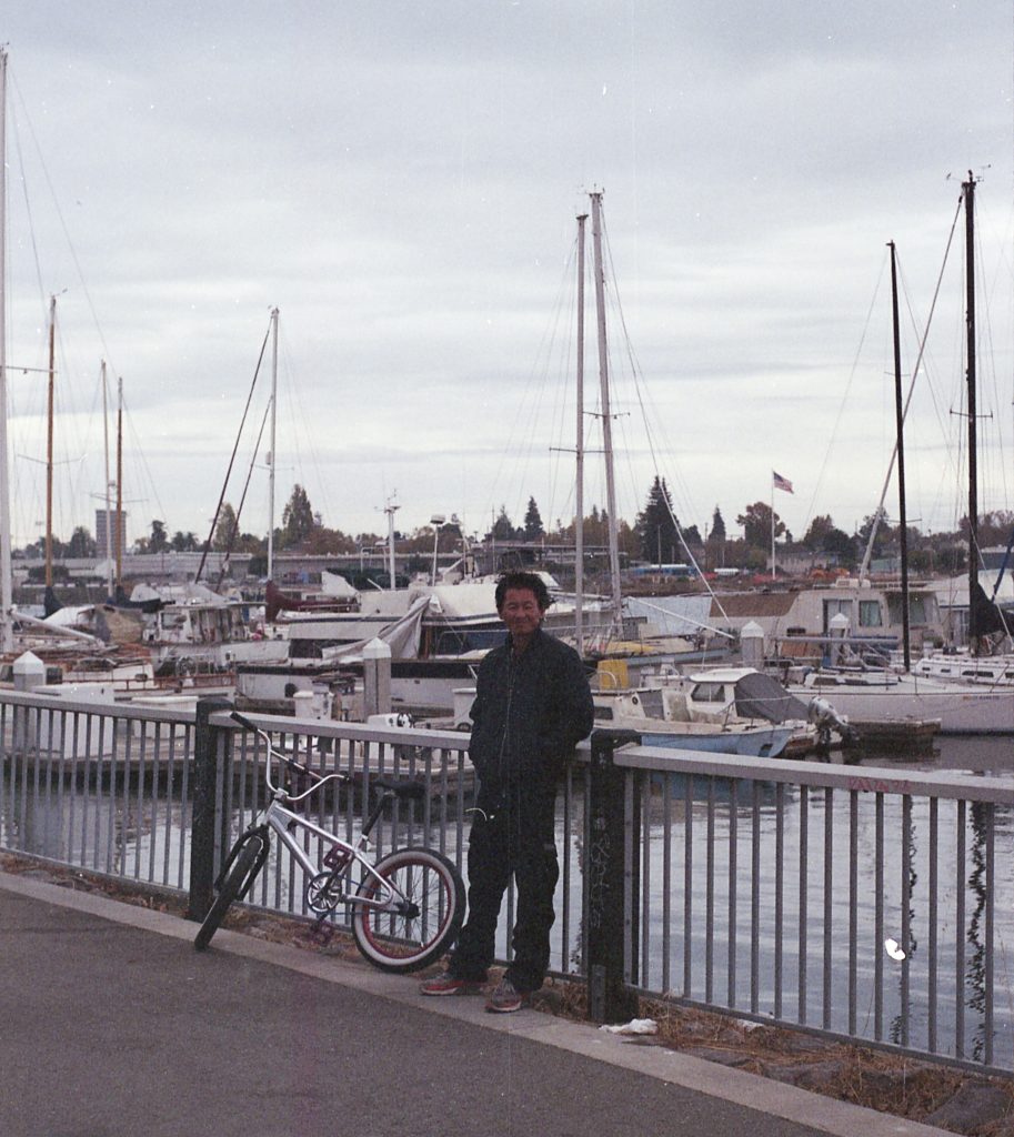 An encampment resident leans against a railing with his bike. Behind him the water of the Marina and sail boats are visible.