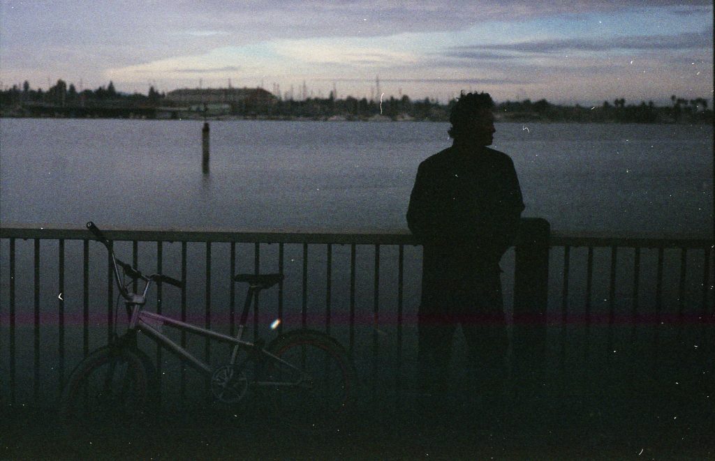 The silhouette of an encampment resident and his bike leaning up against a fence. Behind the resident lies the Marina, with water and sailboats visible.