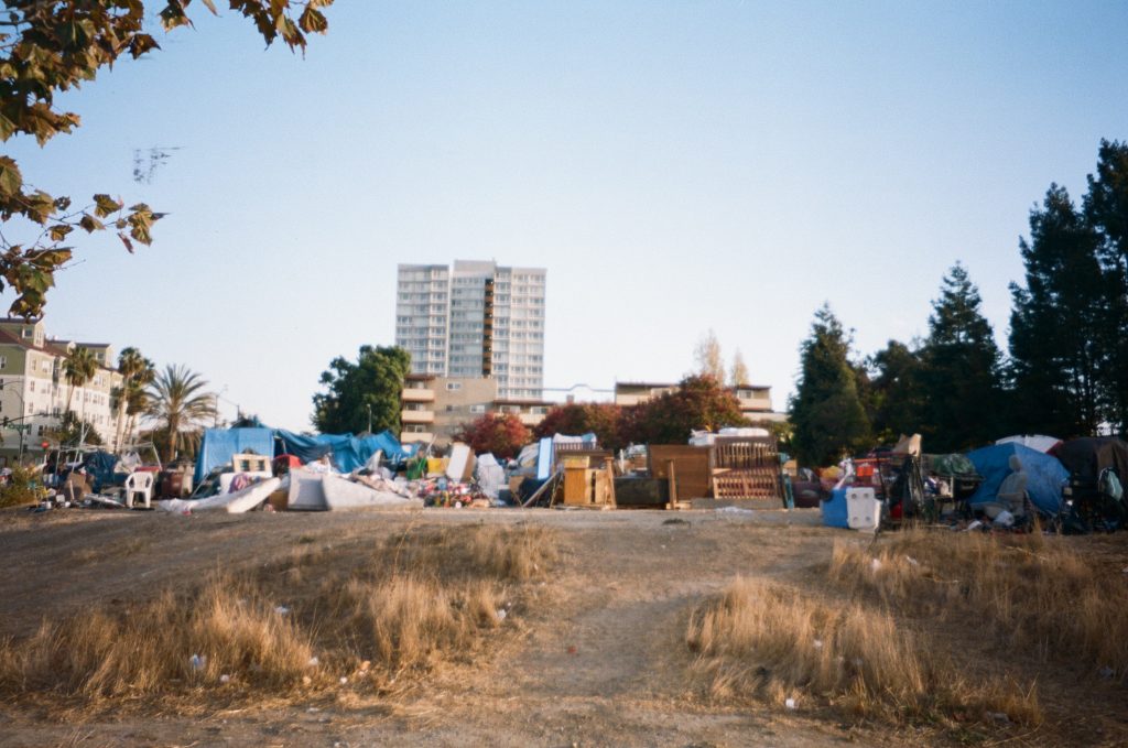 A photo of a homeless encampment surrounded by buildings in Downtown Oakland in the background.