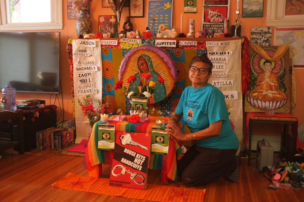 boona cheema sits in her home in front of an altar for lives lost on the street. The altar consists of flowers, photos, and names.