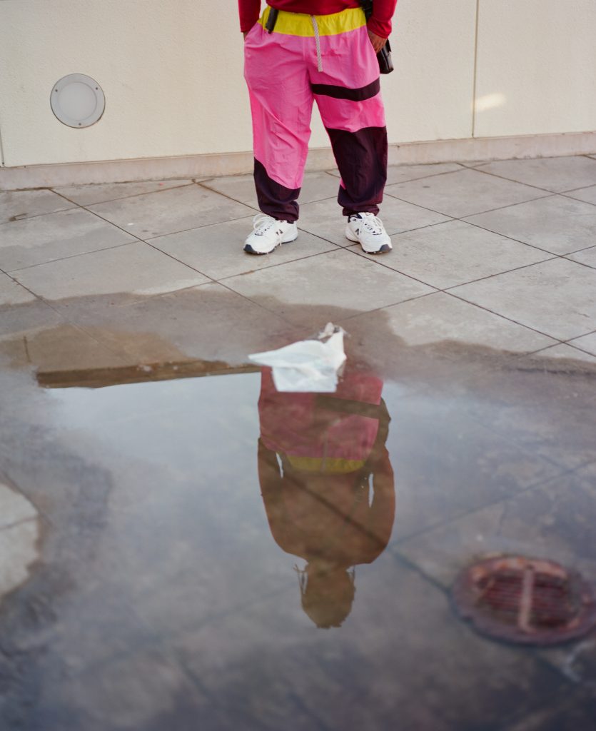 Lil B's reflection in a puddle outside the Cliff House in San Francisco. He is wearing hot pink pants and white sneakers.