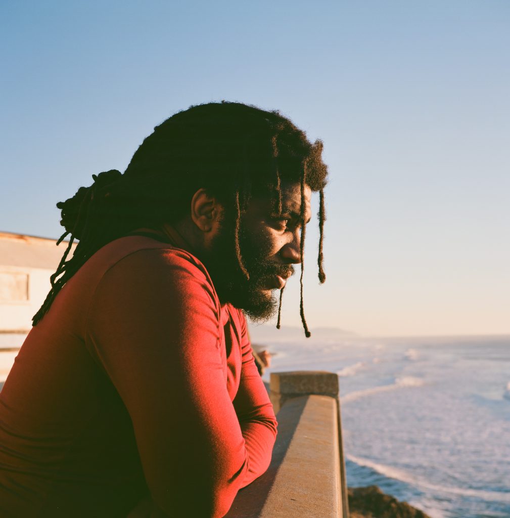 Li B looks out over the ocean. The photo is a profile. You can see his contemplative stare as he looks out over the ocean. Dreadlocks hang in front of his face. His red T-shirt poses a bright contrast against the blue ocean in the background.
