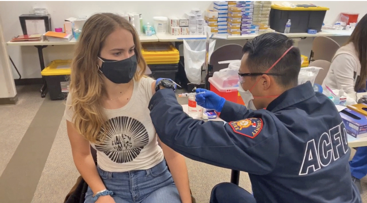 A fire fighter gives the vaccine to a blonde haired woman wearing a black mask.