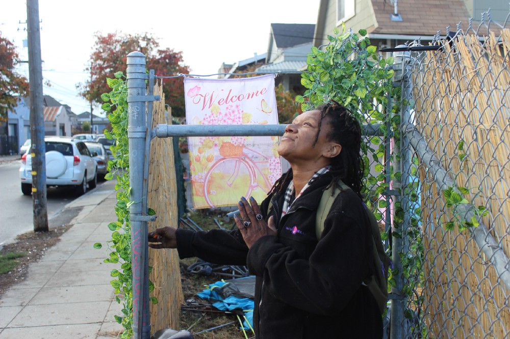 A Black woman stands in front of a chain link gate that has been decorated by a plastic ivy plant that has been woven through the metal fencing. A sign that reads "welcome" hangs on the gate, which is an entrance to the 37MLK homeless encampment. The woman looks up at the sky and holds one hand to her heart, as if in prayer or thanks to a higher being. She has a smile on her face.