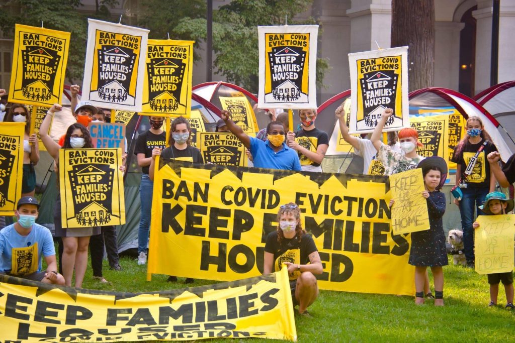 A group of people holding banners and signs that say "ban COVID evictions, keep families housed." People have masks on and some are holding fists in the air. It is a mixed age group containing both young children and adults.