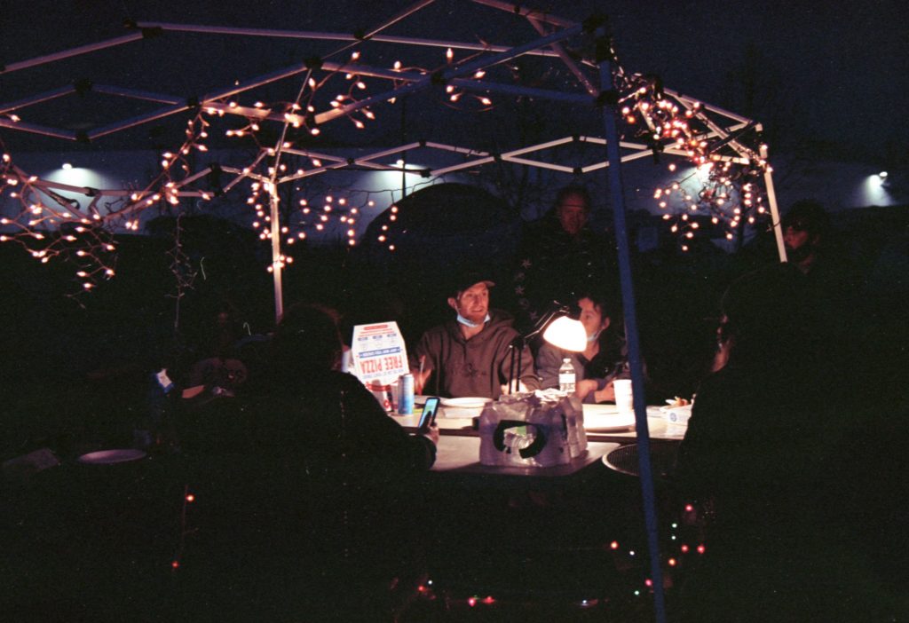 In the dark, a group of encampment residents and their supporters sit under a tent and strategize. Their faces are lit up by a string of twinkle lights hanging from the easy-up above them.