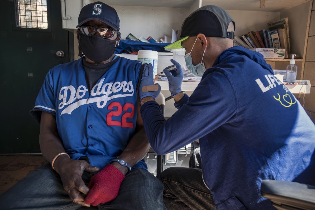 An RN gives the vaccine to a Black man wearing a hat, sunglasses, a Dodgers jersey, and one red glove.