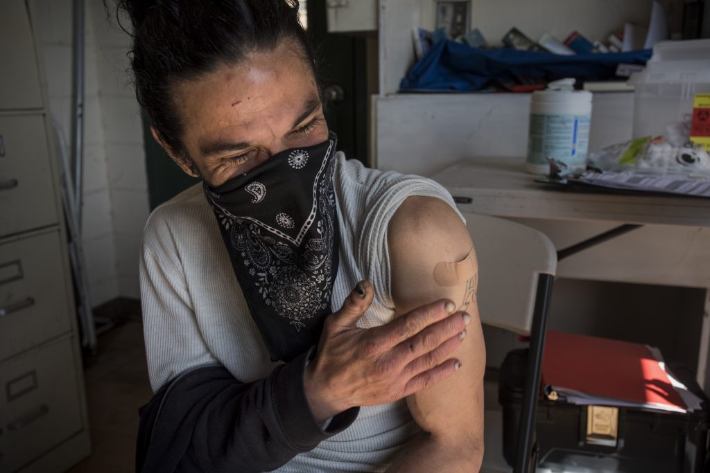 A white man with black hair and a black bandana wrapped around his face as a mask pats his arm. A bandaid can be seen where he just received his COVID vaccine.