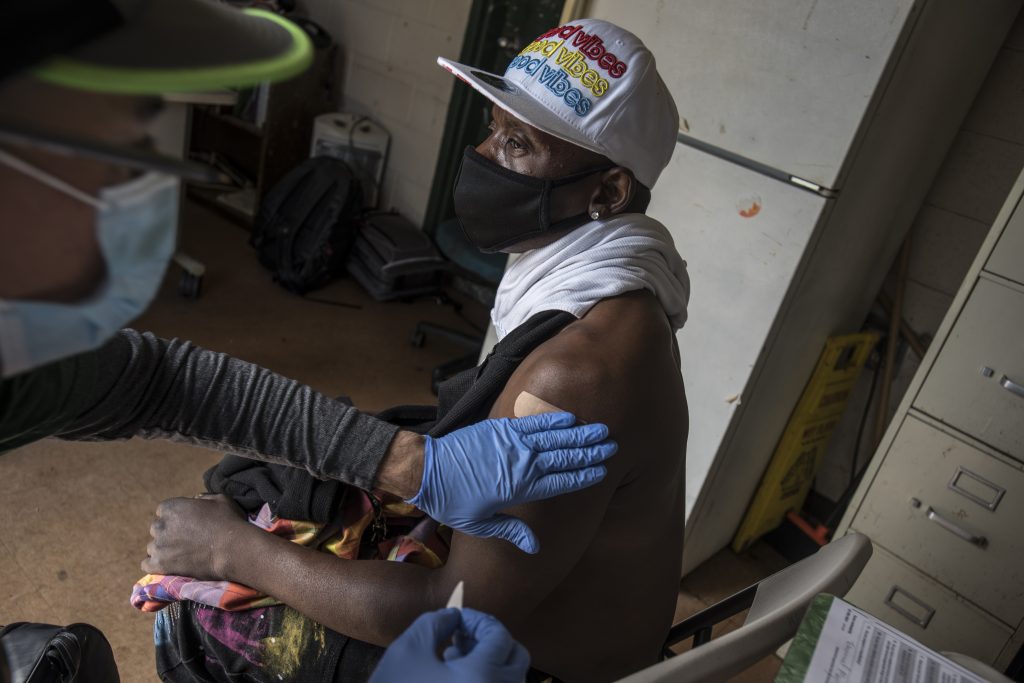 A gloved doctor puts a bandaid on the arm of a Black man in a hat that reads "good vibes" after administering his vaccine.