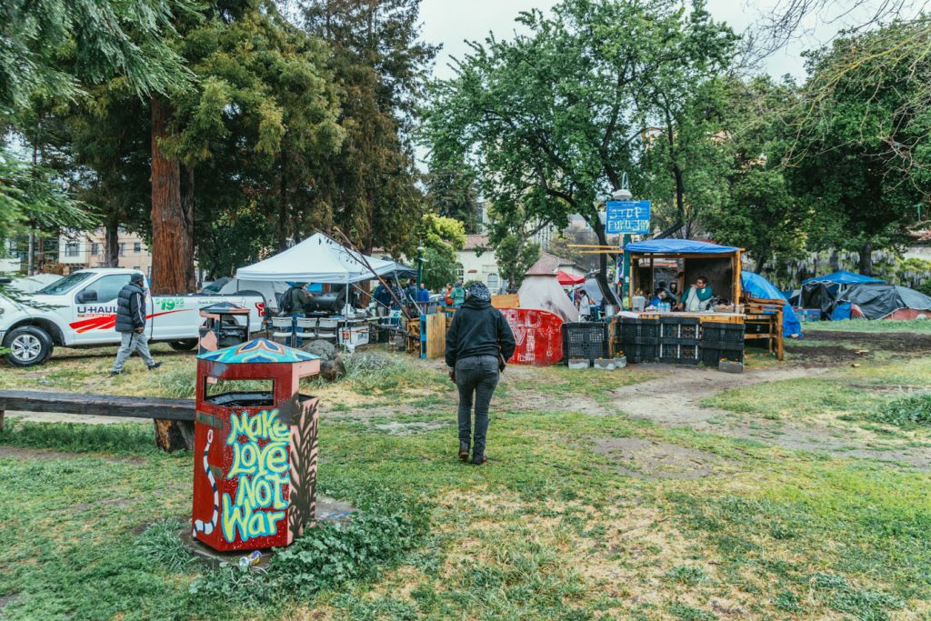 The people's park community kitchen with tents in the background. In the foreground a trash can is painted with a mural that reads "make love not war."